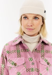 Vrouw met blauwe ogen draagt een crèmekleurige gebreide beanie en een coltrui onder een roze fleecejack met groene en lichtroze zigzagpatronen.