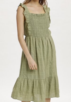 Femme portant une robe midi sans manches vert olive avec un décolleté carré à volants et une jupe froncée, debout sur fond blanc.