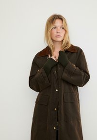 Donna bionda che indossa una giacca oversize marrone scuro con grandi tasche e guanti senza dita verde oliva, in piedi contro uno sfondo bianco.