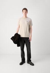 Beige bomulds-T-shirt med et sort logo, parret med mørke jeans og sorte læderstøvler. Modellen holder en sort jakke over den ene arm.