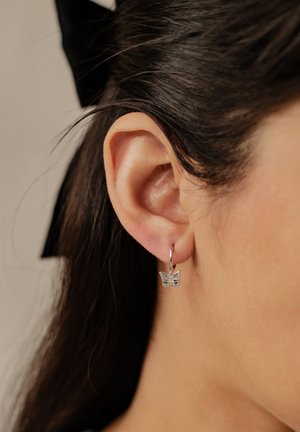 Pendiente de aro en forma de mariposa de plata, con intrincados detalles grabados en la mariposa, colgando de un pequeño aro, mostrado en la oreja de una persona.