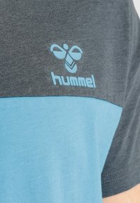 Herret-shirt med en farveblokdesign bestående af mørkegrå og lyseblå sektioner. Foran er der et lyseblåt "hummel" logo.