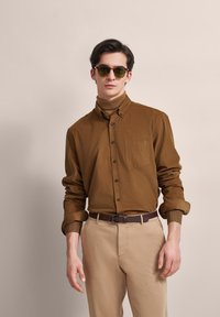 Braunes Hemd mit Knöpfen und Brusttasche, kombiniert mit einem braunen Gürtel und hellbeigen Hosen. Leichtes Material, langärmlig mit hochgerollten Ärmeln, strukturiertem Rollkragen.