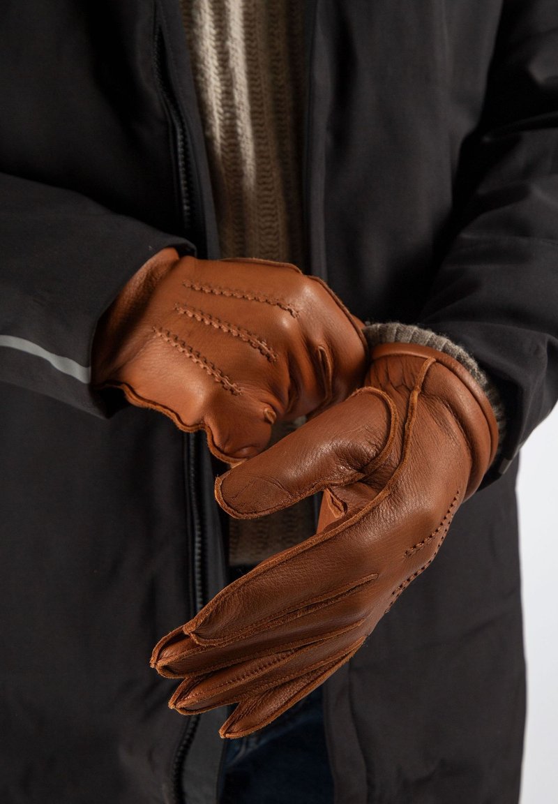 Bruine leren handschoenen met gestikte details, een aansluitende pasvorm en een gestructureerd oppervlak; gedragen door een persoon met een beige trui en een zwart jack.