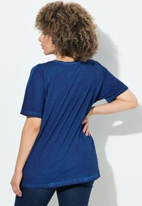 Žena s kudrnatými vlasy, která nosí volné modré tričko s krátkým rukávem a tmavé džíny, stojí zády, s jednou rukou na boku.