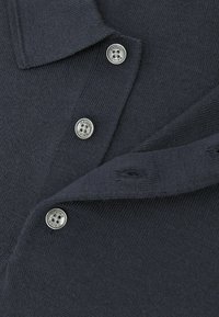 Tumši zils polo krekls no adīta auduma ar apkakli un sudraba pogām ar gravējumu. Satur smalku tekstūru un šuvju detaļas.