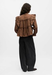 Blouse marron à volants avec des manches courtes, dotée de bords festonnés et d'un dos froncé. Associée à un jean noir large.