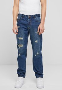 Jeans din denim de culoare bleu deschis cu accente distinse, având rupturi și detalii desfăcute. Design cu picior dreapt și asociat cu un top alb fără mâneci și adidași.