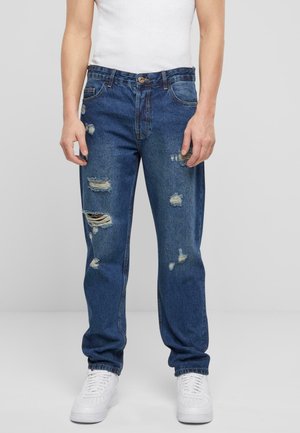 Jean en denim bleu clair avec des accents usés, présentant des déchirures et des détails effilochés. Design à jambe droite associé à un débardeur blanc et des baskets.
