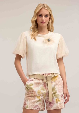 Camiseta crema con mangas cortas y transparentes en forma de puff y un broche decorativo, combinada con pantalones cortos florales rosas que tienen un cinturón y hebilla beige.