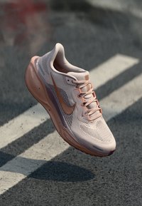 Zapatilla deportiva de color rosa claro con parte superior de malla, cordones y un prominente logo de swoosh. Cuenta con una suela acolchada y acentos texturizados en tonos más oscuros.
