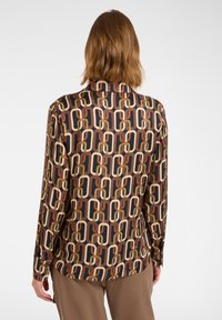 Person with shoulder-length hair wears long-sleeve shirt with brown, beige, and black linked chain pattern, paired with light brown pants.