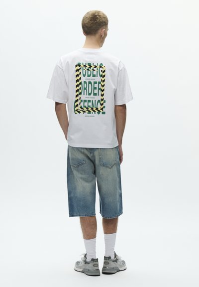 White cotton t-shirt with graphic print featuring green text and yellow-black striped frame on the back. Worn with light blue denim shorts.