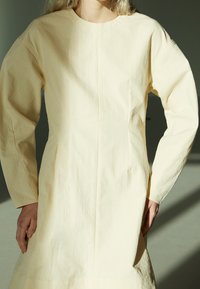 Vestido de color crema hecho de una tela texturizada, con mangas largas, escote redondo y bolsillos laterales. Silueta sencilla y recta.