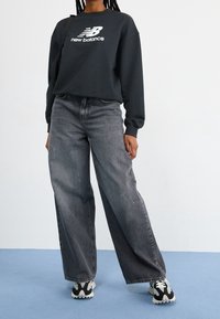 Felpa nera con logo "New Balance" bianco, abbinata a jeans grigi a gamba larga e sneakers in bianco e nero con un design a motivi.