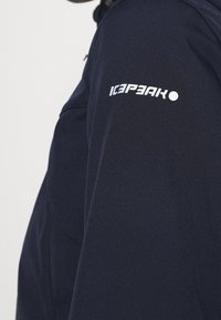 Tummansininen pehmeäkuorijakku, jossa on sileä pinta, valkoinen logo vasemmassa hihassa ja muotoiltu malli.