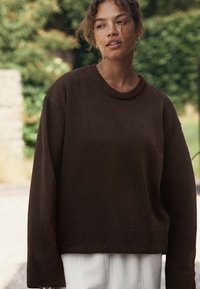 Ruskea neulepaita pyöreällä pääntiellä ja löysillä, pitkillä hihoilla. Materiaali vaikuttaa pehmeältä ja istuvuus on hieman väljä.