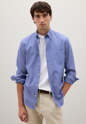 Blue and white striped long-sleeve shirt with button collar, rolled sleeves, and a chest pocket, worn over a white t-shirt and beige trousers.