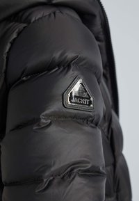 Svart pufferjacka med vadderad textur, dragkedjestängning och en triangulär silverlogotyp på vänster arm. Slät tyg.