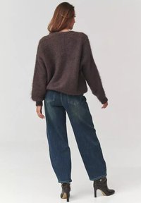 Brązowy, puszysty sweter o luźnym kroju, zestawiony z niebieskimi dżinsami z wysokim stanem. Modelka nosi czarne botki za kostkę na wysokim obcasie.
