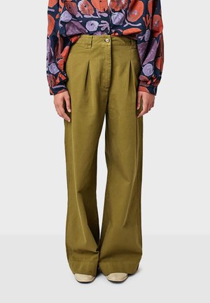 Pantaloni a gamba larga in cotone verde oliva con pieghe frontali, rifiniti con una chiusura a singolo bottone. Indossati con una camicia a maniche lunghe a fantasia.