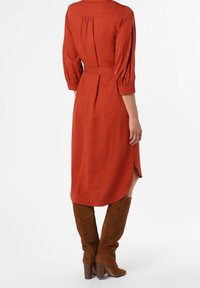 Robe couleur rouille avec une taille ceinturée, des manches trois-quarts et des fentes latérales ; associée à des bottes hauteur genou en suède beige et un talon épais.