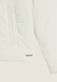 Chaqueta de tela blanca con acanalado y un bolsillo en el pecho, que presenta una pequeña etiqueta de marca en tono dorado, acentuando su diseño texturizado.