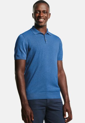 Lachende man die een blauw poloshirt met korte mouwen en donkere broek draagt, staand tegen een effen witte achtergrond.