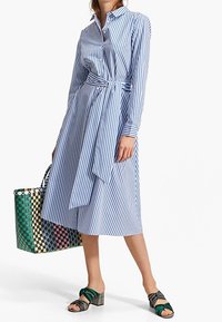 Raidallinen mekko vaaleansinisena ja valkoisena, vyötäröllä vyö ja pitkät hihat. Kudottu kassi vihreänä, vaaleanpunaisena ja mustana. Mustat sandaalit.