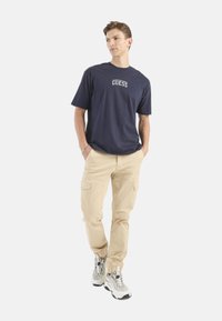 Camiseta de algodón azul marino con el logo "GUESS", combinada con pantalones cargo beige y zapatillas con estampado; ajuste casual y diseño relajado.