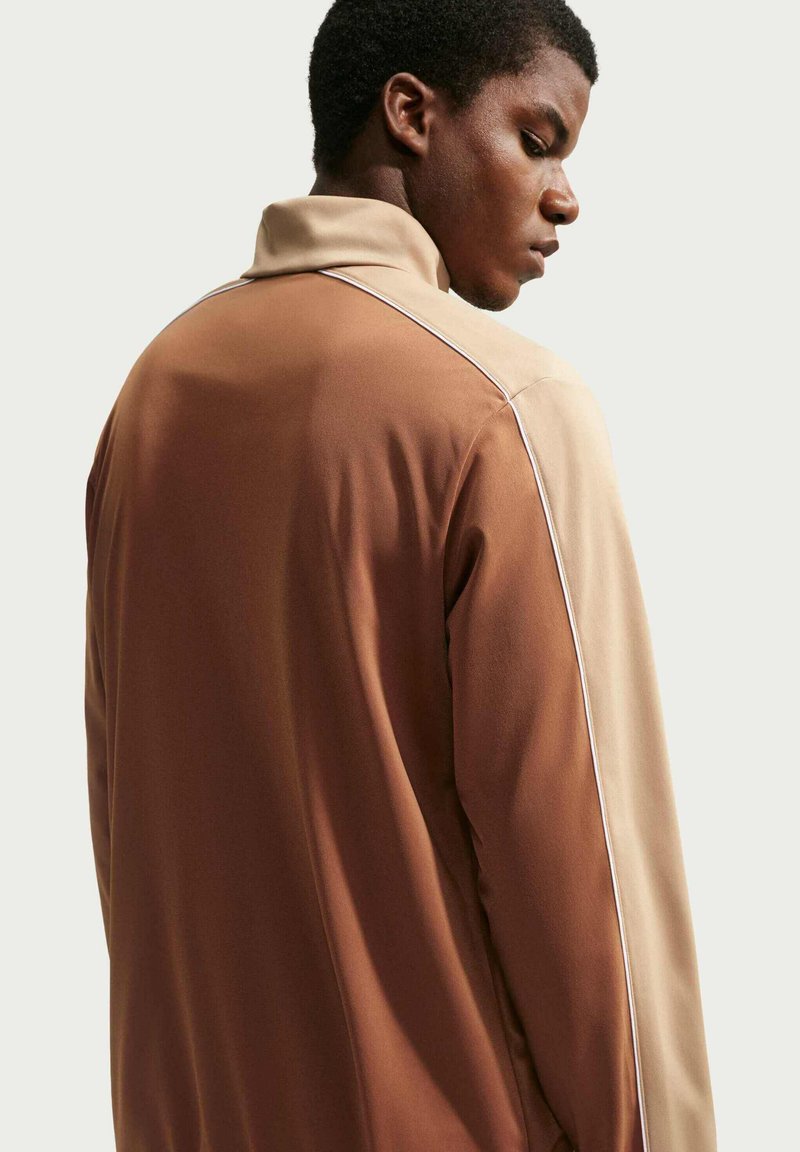 Ung mand ser ned, iført en tofarvet brun og beige jakke med høj krave mod en ensartet lys baggrund.