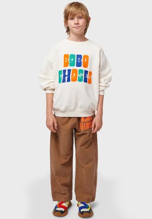Felpa bianca con il testo colorato "BOBO CHOSES", abbinata a pantaloni in corduroy marroni con stampa del logo arancione e scarpe multicolori.