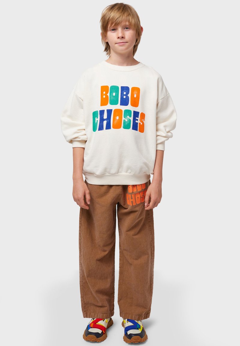 Sudadera blanca con el texto "BOBO CHOSES" colorido, combinada con pantalones de pana marrones que presentan un logo en naranja y zapatos multicolores.