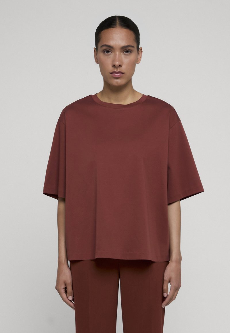Femme portant un haut ample à manches courtes rouge foncé et un pantalon assorti, face à un fond uni gris clair.