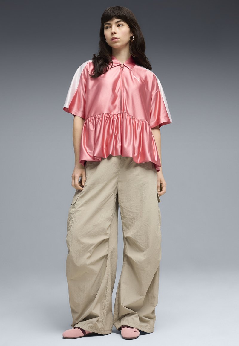 Jeune femme aux cheveux longs et foncés portant un chemisier rose brillant à manches courtes, un pantalon large beige et des chaussures roses, debout devant un fond gris.