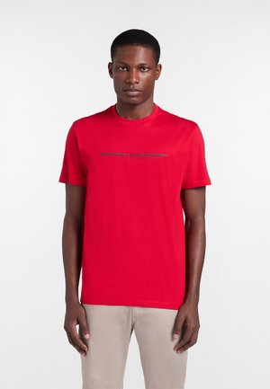 Muž v červenom tričku s krátkym rukávom s malým stredovým grafickým textom, v svetlo béžových nohaviciach, stojaci pred jednoduchým pozadím.