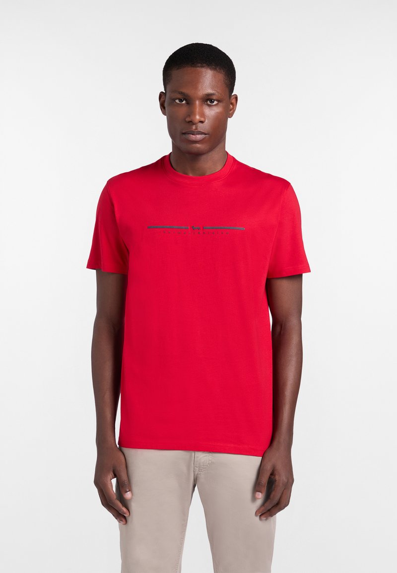 Hombre con camiseta roja de manga corta con un pequeño texto gráfico en el centro, combinado con pantalones beige claro, de pie frente a un fondo liso.