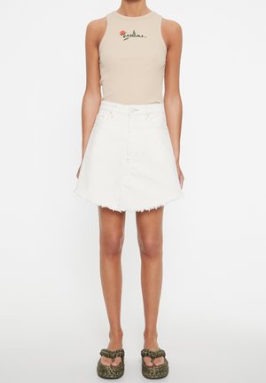 Femme portant un haut beige sans manches à motif floral, une jupe en jean blanche effilochée et des sandales à plateforme vertes tressées sur fond blanc.