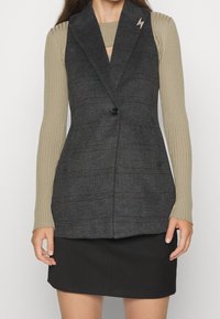 Gilet grigio su misura con rever a punta, chiusura con un singolo bottone nero e un sottile motivo a quadri, abbinato a un top a maniche lunghe a coste e una gonna nera.