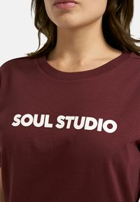 Burgundy cotton t-shirt featuring a white "SOUL STUDIO" graphic print at the chest, with a crew neck and short sleeves.