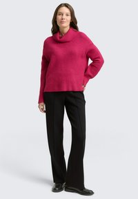 Fuchsiastrickpullover mit einem Schalkragen und langen Ärmeln, kombiniert mit schwarzen, geraden Hosen und schwarzen Schuhen vor einem hellen Hintergrund.