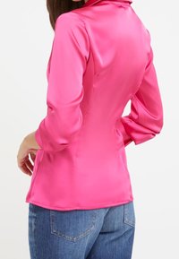 Blouse rose en satin ajustée avec des manches longues et un col rabattu, présentant une texture lisse et un ourlet arrondi, associée à un jean bleu.