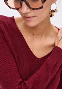 Burgundowy sweter z dekoltem w serek, prążkowaną fakturą i długimi rękawami. Akcesoria to duże okulary przeciwsłoneczne w stylu tortoise i wyrazista kolczyk.