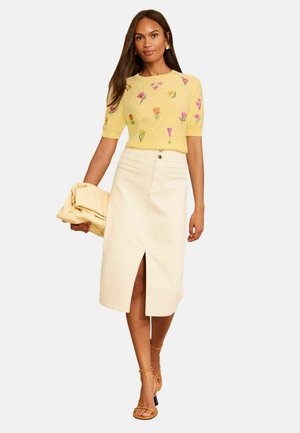 Woman wearing a yellow floral embroidered short-sleeve top, cream midi skirt with front slit, and tan strappy heeled sandals, holding a folded jacket.