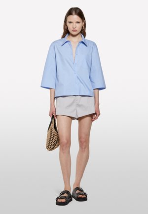 Chemise bleu clair à manches courtes avec boutons, détail de poche, associée à un short gris, des sandales noires et un sac en osier.