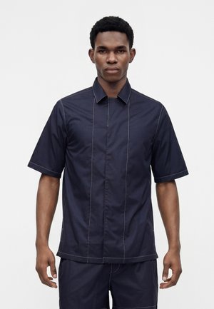 Homme debout, face à l'avant, portant une chemise bleu marine foncé à manches courtes boutonnée et un short assorti sur un fond blanc uni.