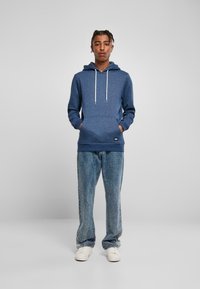 Felpa cappuccio blu scuro con cordoncino, tessuto strutturato, tasca a marsupio, abbinata a jeans chiari strappati e sneakers bianche.
