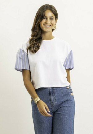 Jeune femme aux longs cheveux bruns souriante, portant un haut blanc avec des manches à rayures bleues, un jean taille haute bleu, ainsi qu'un bracelet et des boucles d'oreilles en or.