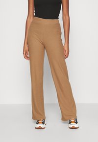 Pantalon large côtelé de couleur brun chaud ; taille élastique ; associé à des baskets noires et blanches. Texture lisse, design décontracté.