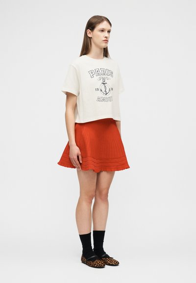 Νέα γυναίκα που φοράει λευκό κομψό κοντό μπλουζάκι με σχέδιο άγκυρας, πορτοκαλί πλισέ φούστα, μαύρες κάλτσες και παπούτσια με λεοπάρ σχέδιο, στέκεται πλευρικά.